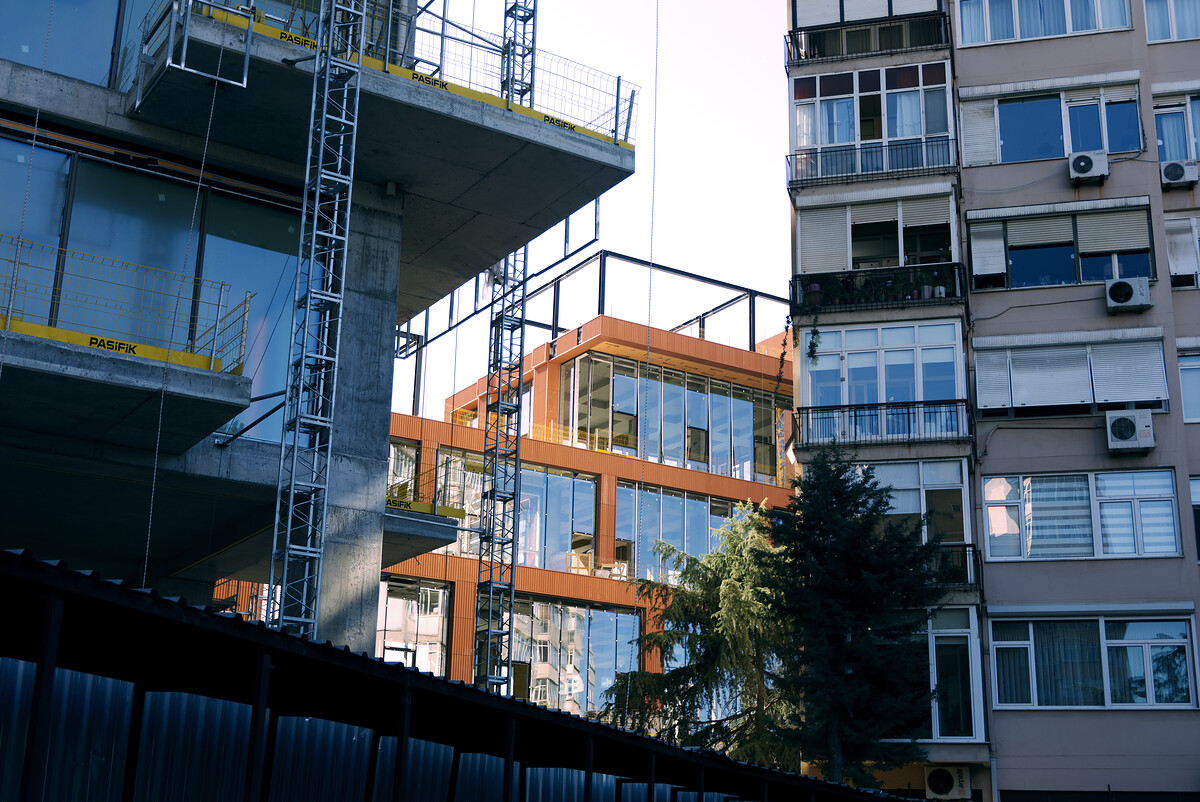 A concrete building under construction beside an orange-framed glass building and an apartment block, with a pine tree in front